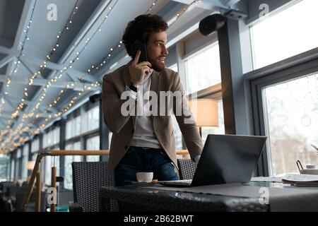 Angenehmer intelligenter Manager im Gespräch mit Kollegen, Partner mit Smartphone, Blick beiseite, am Fenster. Stockfoto