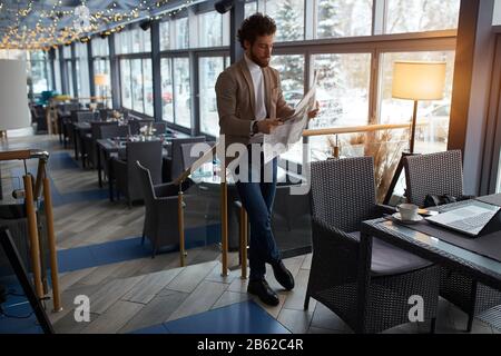 Eleganter Kerl, der mit gekreuzten Beinen steht und Spaß am Lesen von Zeitung, Hobby, Interesse, Massenmedien hat. Foto in voller Länge Stockfoto