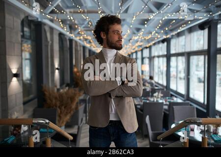 Erfolgreicher Kleinunternehmerbesitzer, der mit verschränkten Armen in seinem Café steht, beiseite schaut, weg, am Fenster, Nahaufnahme Foto. Erfolg Stockfoto