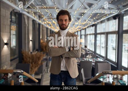 Der junge, lächelnde bärtige Mann hat ein neues Café, ein Restaurant, ein Nahfoto eröffnet. Willkommen in einem neuen Café mit modernem Interieur. Stockfoto