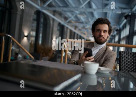 Junger gutaussehender Mann, der sein Smartphone benutzt, auf dem Stuhl im Café sitzt, unscharfen Vordergrund, Mann wartet auf Partner, Kunden. Nahaufnahme Stockfoto
