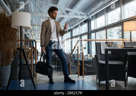 Junger eleganter Mann, der im Café nach Wi-Fi-Verbindung sucht und auf Freundin, Kunden und Kunden wartet. Foto in voller Länge Stockfoto