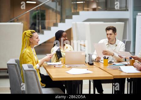 Junger attraktiver Mann, der seinen Kollegen einen Bericht vorliest, zwei angenehme Frauen lächeln, ihn betrachten, Spaß im Büro haben.Nahaufnahme Foto Stockfoto