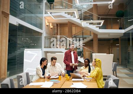 Junger kluger Mann, der seine Kollegen während des Treffens berät. Gutaussehender Mann in rotem Hemd, der seinen Freunden drinnen Empfehlungen gibt. Stockfoto
