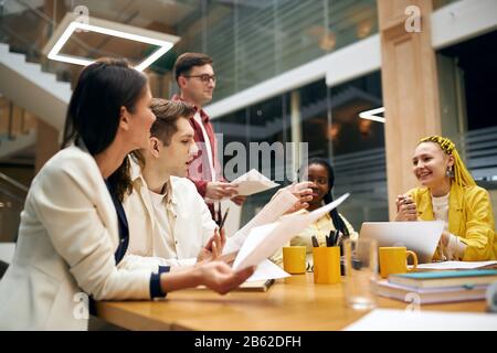Junge Leute, die Dokumente analisieren, während sie am Schreibtisch sitzen, kreativer und innovativer Designer, der ein Projekt abhält und es den Partnern vorstellt Stockfoto