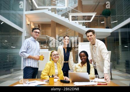 Erfolgreiches, ambitioniertes Business Team am Arbeitsplatz: Junge, fröhliche Leute, die Kamera, Job, Beruf, ccupation, Glaswände und Fenster betrachten Stockfoto