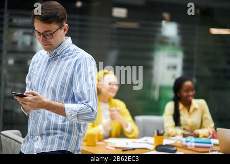 Der junge ehrfürchtige Mann hat sich verliebt, kann sich nicht auf die Arbeit konzentrieren, ein fröhlicher lächelnder Kerl telefoniert, SMS an Partner, Freundin Stockfoto