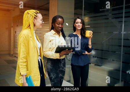 Selbstbewusste lächelnde afro-Geschäftsfrauen, die Notizblock halten, Notizen machen, während sie mit ihren Pfartnern sprechen, writimg irgendwo auf dem Papier. Naht Seite wetteifern Stockfoto