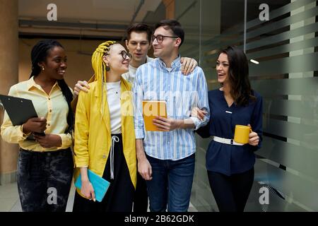 Fröhliche Gesellschaft junger Menschen, die durch den Koridor spazieren, positive Gefühle und Emotionen ausdrücken, freie Zeit haben, sich nach Geschäftsmetin ausruhen Stockfoto