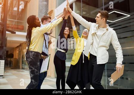 Positive aktive junge Mitarbeiter heben gemeinsam ihre Arme auf und zeigen ihre Einheit, Zusammengehörigkeit, eine für alle, alle für eine, Teambuilding Stockfoto