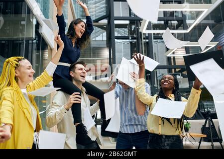 Kreativ freundliche Mitarbeiter, die im Büro hängen, Angestellte, die Dokumente, Papiere, schreiende, brünette Frau werfen, die auf den Scholidern ihres c sitzt Stockfoto