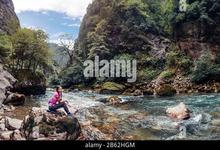 Süße Junge Frau, die auf dem Stone am Ufer des schnellen Gebirges sitzt. Hikerin in Sportlicher Kleidung im felsigen Canyon Stockfoto