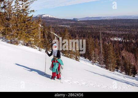 Eine in Rot gekleidete Klettererin steigt einen verschneiten Berghang hinauf. Winter Clear Sky Day. Im Hintergrund Ural-Gebirge, russland Stockfoto