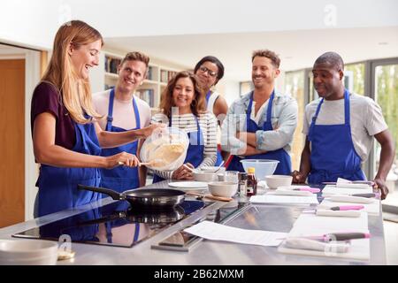 Weibliche Lehrerin, Die Pancake Auf Dem Herd In Der Kocherklasse Macht, Während Erwachsene Schüler Darauf Achten Stockfoto