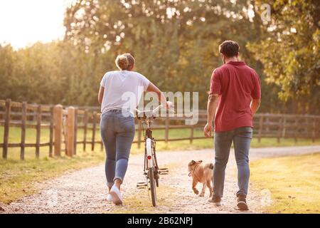 Rückansicht Des Paares Mit Haustierhund Mit Dem Push Bike Auf Der Country Lane Bei Sonnenuntergang Stockfoto