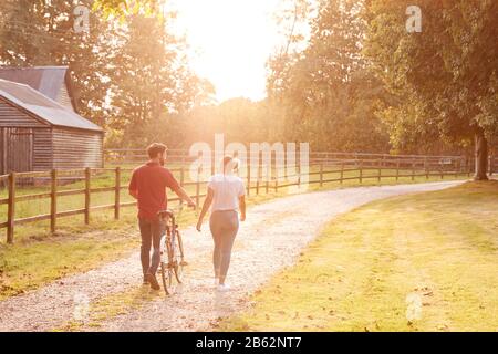 Rückansicht Eines Romantischen Paares Bei Sonnenuntergang, Das Mit Dem Fahrrad Entlang Der Country Lane Spazieren Geht Stockfoto
