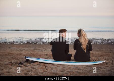 Rückansicht Eines Nachdenklichen Paares Mit Wetsuits Auf Surfstaycation Mit Blick Auf Die Wellen Zum Meer Stockfoto