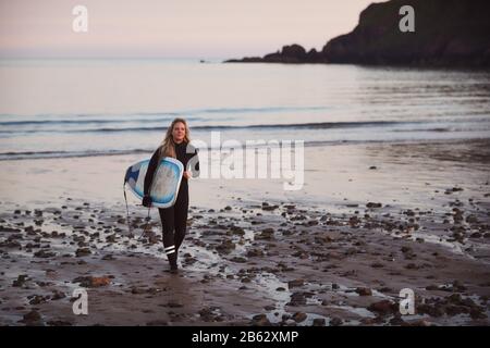 Frau, Die Surfbrett Mit Wetsuit Trägt, Während Sie Aus Dem Meer Läuft Stockfoto