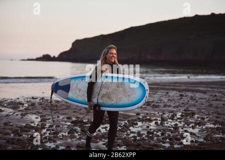 Frau, Die Surfbrett Mit Wetsuit Trägt, Während Sie Aus Dem Meer Läuft Stockfoto
