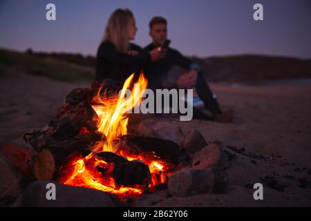 Ein Paar Sitzt Auf Surfboard Von Camp Fire On Beach Und Nutzt Das Handy, Während Sun Hinter Ihnen Sitzt Stockfoto