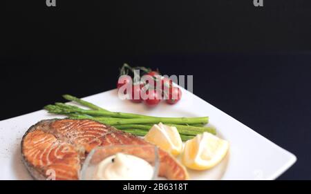 Lachssteaks mit Gemüse und Gewürzen: Spargel, Tomaten, Ingwer, Pfefferkörner, Salz, Chili, Zwiebel, Zitrone und Olivenöl auf schwarzem Grund. Heilen Stockfoto