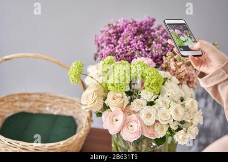 Eine Frau macht ein Foto auf ihrem Telefon mit Blumen. Blumengeschäft Konzept . Blumenfrau schafft Blumenarrangement in einem Korbkorb. . Blumen Lieferung. Stockfoto