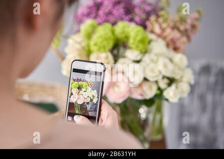 Eine Frau macht ein Foto auf ihrem Telefon mit Blumen. Blumengeschäft Konzept . Blumenfrau schafft Blumenarrangement in einem Korbkorb. . Blumen Lieferung. Stockfoto