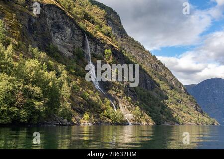 Wasserfall fällt, norwegischer Fjord Aurlandsfjord Spiegelreflexion. Heller Blick auf die wilden Berge in der Natur Norwegens. Sonniger Tag in Skandinavien. Stockfoto