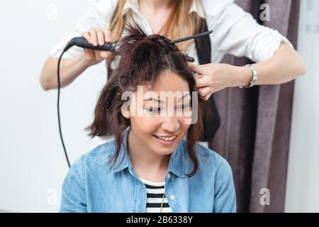 Frau Friseur machen Frisur mit Lockenstab für langes Haar der jungen Frau mit Smartphone im Beauty-salon Stockfoto