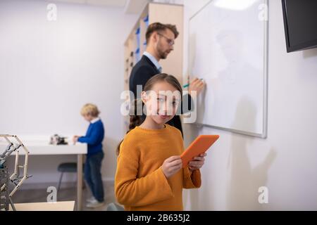 Männliche Lehrerin schreibt auf Whiteboard, Mädchen hält Tablette, Junge steht am Schreibtisch Stockfoto