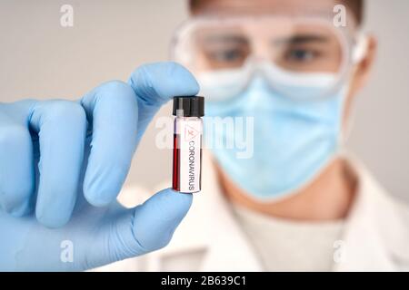 Selektiver Fokus auf Reagenzglasbehälter mit Blutprobe in der Hand eines selbstbewussten Arztes, mit Schutzanzug und Gesichtsmaske, die gegen Unschärfe steht Stockfoto