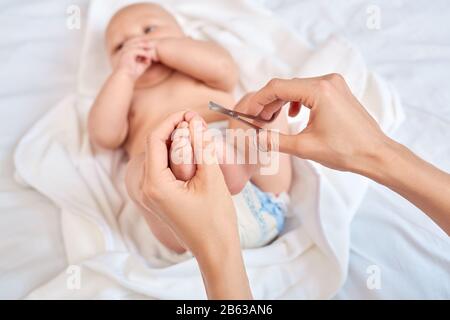 Junge Mutter kümmert sich um den kleinen Sohn, der Einwegwindeln trägt, die den Daumen auf dem Bett liegen, zu Hause schneiden Nägel auf Zehen mit kleiner Schere c Stockfoto
