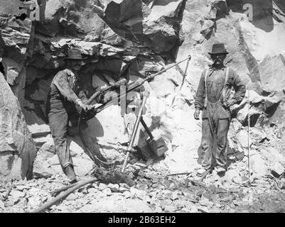 Zwei Männer, die eine Dampfbohrmaschine c.-210 betreiben. Sie bohren ein Loch bis zu einer gewissen Tiefe in die Steinmauer. Der Mann auf der rechten Seite hat einen schmalen Stab, der in das Loch eingeführt werden kann, um die Tiefe des Lochs zu überprüfen. Wenn das Loch tief genug ist, wird Dynamit in das Loch gestaut, bis die Mannschaft bereit ist, die Felswand wegzusprengen, um Platz für eine neue Eisenbahn oder Straße zu schaffen oder usw., um meine anderen Vintage-Bilder zu sehen, Suche: Prestor Vintage Work Stockfoto