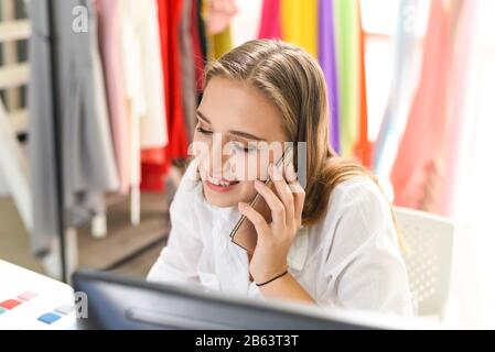 Junge Frau, die am Schreibtisch mit Computer-pc arbeitet und Smartphone oder Telefon Mit Jemandem auf ihrem Handy über das Geschäft an ihrem Arbeitsplatz Spricht Stockfoto