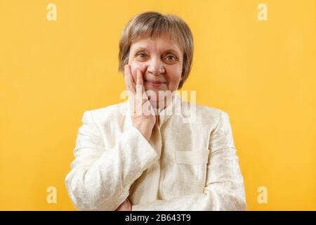 Porträt einer lächelnden grauhaarigen Seniorin mit Handfläche, die Wange auf gelbem Hintergrund hält. Stockfoto