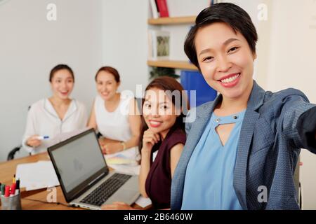 Fröhliche junge vietnamesische Unternehmerin fotografieren mit ihren lächelnden Kollegen nach einem erfolgreichen Treffen Stockfoto