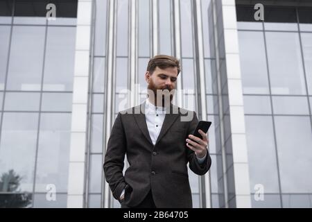 Junger bärtiger Geschäftsmann hält Handy mit App SMS-Nachricht per Jacke im Freien. Erfolgreicher Unternehmer in Form gekleidet Stockfoto