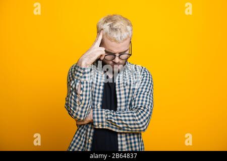 Der kaukasische Mann mit blondem Haar und Bart berührt seinen Kopf, während er über etwas nachdenkt und auf einem gelben Hintergrund posiert Stockfoto