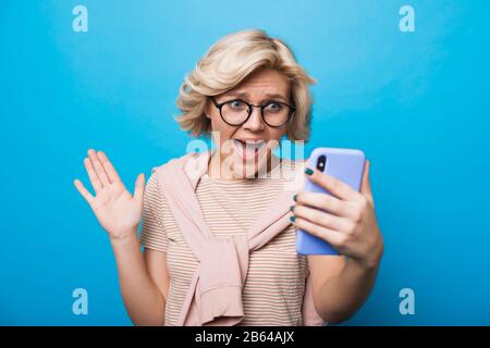 Charmante kaukasische Frau mit blondem Haar ist von etwas überrascht, während sie ein Telefon hält und auf einem blauen Hintergrund posiert Stockfoto