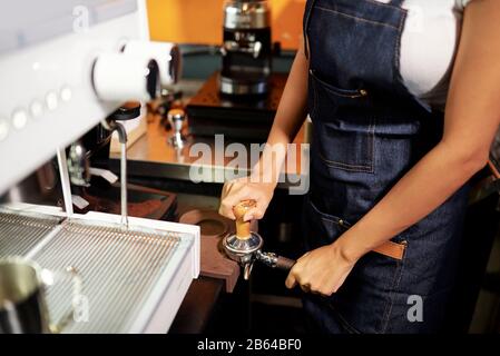 Nahaufnahme des weiblichen Baristas, der gemahlenen Kaffee im Auslauf-Portafilter eintampelt, bevor er mit der Espressomaschine gebrüht wird Stockfoto
