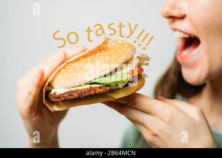 Das Mädchen hält einen Hamburger mit den Händen und isst ihn mit Appetit. Die Aufschrift ist so lecker. Nahaufnahme. Stockfoto