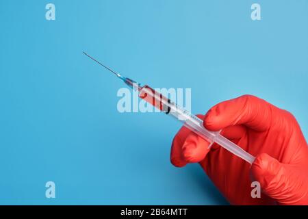 Doktor in roten Handschuhen hält eine Spritze mit einem Impfstoff auf blauem Grund. Nahaufnahme. Stockfoto