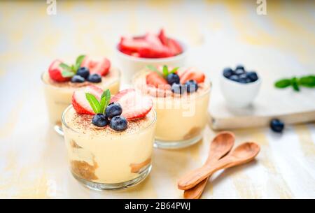 Hausgemachte, exquisite Dessert Tiramisu in Gläsern dekoriert mit Erdbeere, Heidelbeere, Minze auf weissem Holztisch, Stockfoto