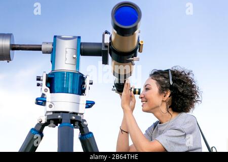 Junge lächelnde Frau, die durch astronomisches Teleskop in die Höhe schaute Stockfoto