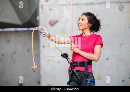 Porträt der schönen Frau Rock-Kletterer, die einen weiteren Kletterer mit Seil hinaufzögert. Künstliche Kletterwand und Ausrüstung im Innenbereich. Stockfoto