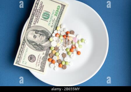 Medikamente Kosten Geld. Drogen und Dollar. Weiße Platte mit mehrfarbigen Pillen und Kapseln und einer 100-Dollar-Rechnung auf einem klassischen blauen Hintergrund. Pillen Stockfoto