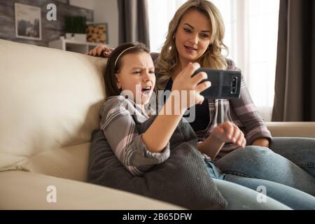 Süßes kleines Mädchen schockiert, nachdem sie ein beunruhigendes Online-Video auf dem Handy neben ihrer Mutter gesehen hat. Moderne Mutter. Stockfoto