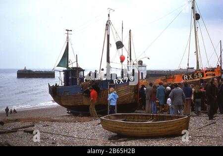 1970er Jahre, historisch, Menschen warten mit alten hölzernen Fischerbooten am Steinstrand von Rock-a-Nore in Hastings, East Sussex, England, Großbritannien, die blau bemalte RX73 und orange bemalte RX58. Die Küstenstadt zählt zu den ältesten Fischerhäfen Großbritanniens und beherbergt die berühmte Hastings Fishing Fleet, die älteste und größte vom Strand ausgehende Fischereiflotte Großbritanniens. Stockfoto