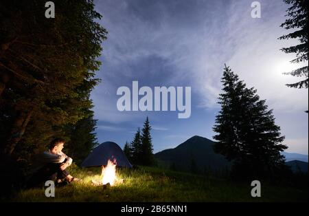 Guy sitzt am Feuer auf dem Hintergrund eines Zelts und eines Fichtenwaldes, der abends einen magischen Blick auf die Berge genießt. Tal der Hügel unter einem Sternenhimmel mit leichten Wolken nach Sonnenuntergang. Stockfoto