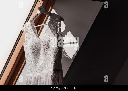 Weißes Hochzeitskleid hängt vor der Zeremonie an den Schultern in der Nähe des Fensters Stockfoto